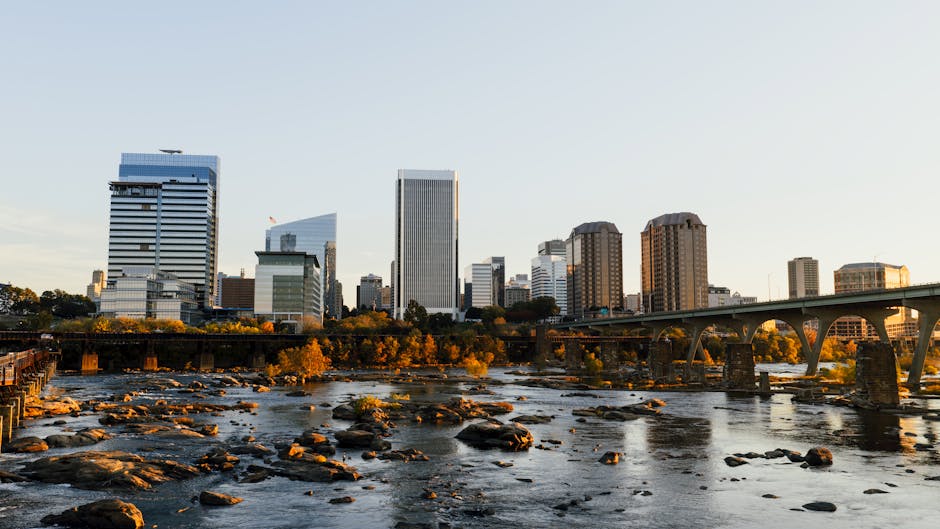 A panoramic view of a city skyline during late afternoon or early evening, featuring modern high-rise office buildings and residential towers along the riverbank. The river in the foreground has visible rocks and small rapids, with some areas reflecting the warm, golden light of the setting sun. A bridge with arched supports spans across the river, connecting different parts of the city. In the background, there are lush trees with autumn foliage along the riverbank, and the sky above is clear with soft lighting. This scene captures an urban environment that could be part of a house removal or moving project, illustrating the transition between residential or commercial properties and transport routes such as bridges and rivers. The overall atmosphere is calm, with natural lighting highlighting the details of both the city’s architecture and the natural landscape.