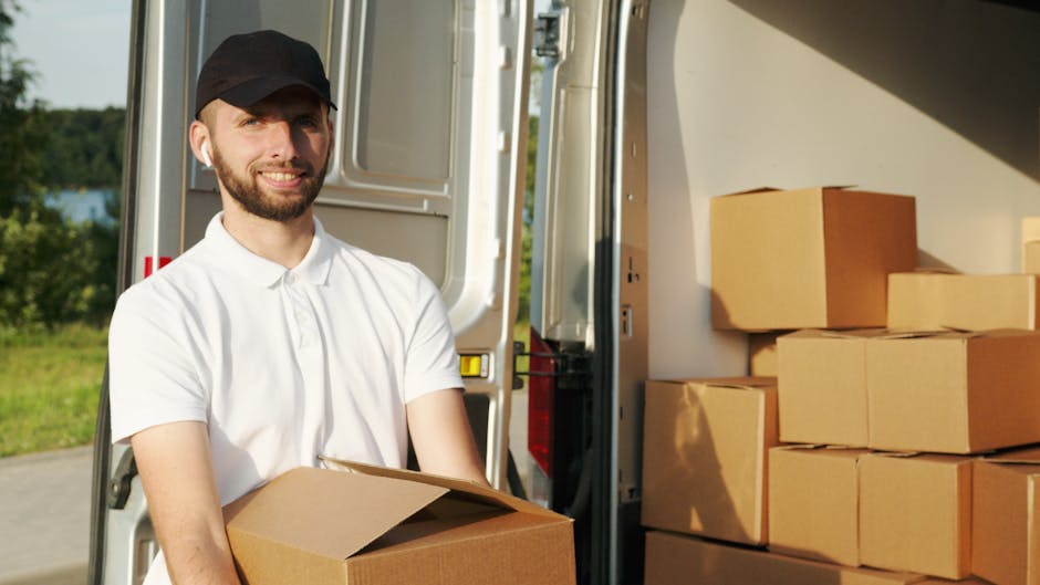 A man with a beard and wearing a white polo shirt and a black cap is standing outdoors in front of an open moving van, holding a cardboard box. The vehicle is positioned on a paved area with green tre