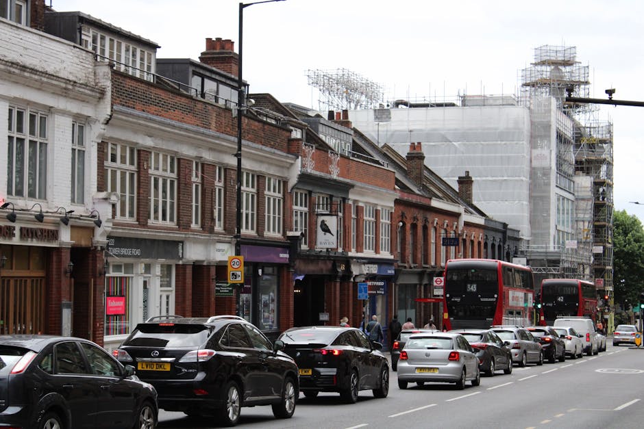 A street scene in East Sheen showing a row of traditional brick-front shops and residential buildings, with some storefronts featuring large windows and signage. Several vehicles, including a black Mercedes-Benz and a white car, are parked along the curb while additional cars are seen moving or waiting in traffic. Two red double-decker buses are captured in the process of approaching or stopping at the bus stop, with some pedestrians walking on the sidewalk. In the background, there is ongoing construction work with scaffolding and white sheeting covering a building, indicating site renovation or development. The scene is shot during daytime under overcast weather, highlighting the typical urban environment where house removals or furniture transport could be taking place, and occasionally, [COMPANY_NAME]'s moving services might be involved in Jon relocations within the area.