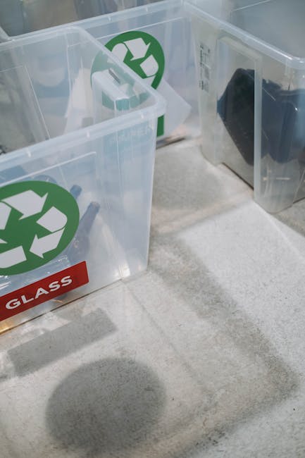 A close-up view of several transparent plastic recycling bins positioned on a light-colored carpeted floor inside a property. Each bin features a green recycling symbol on the front, with one bin labeled 'GLASS' in red. The bins are empty but appear ready for collection of recyclable materials as part of a home relocation or packing and moving process. In the background, there is a glimpse of a doorframe or doorway, indicating the bins are situated near an entrance or within a hallway. The lighting is natural and evenly distributed, highlighting the clean and organized environment. This setup suggests an emphasis on environmentally conscious disposal options supported by Man with Van East Sheen during furniture transport or packing tasks associated with house removals and eco-friendly recycling efforts in East Sheen.