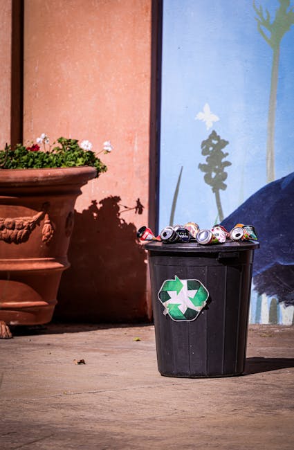 A black plastic recycling bin with a green and white recycling symbol on the front is positioned outside a building, close to a large terracotta flowerpot filled with blooming pink and white flowers. Several crushed aluminium cans are visibly stacked inside and spilling over the top of the bin. The background features a colorful mural painted on the building's wall, depicting a stylized tree and flowers, with the scene illuminated by natural daylight. The setting suggests an outdoor area in East Sheen, where house removals and disposal services such as those provided by Man with Van East Sheen might be involved in the safe recycling or donation of household items during a home relocation or packing process.