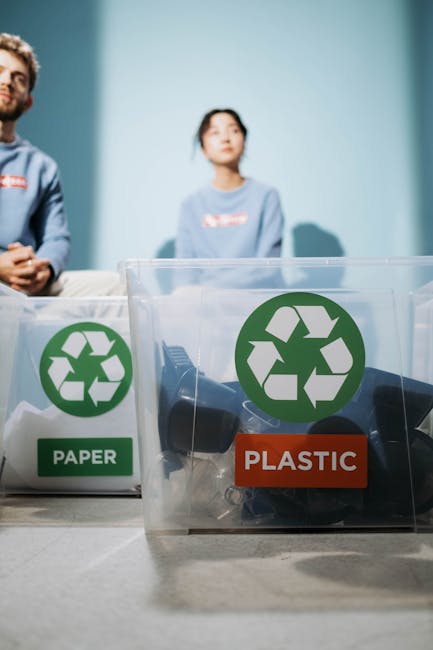 A close-up view of several transparent plastic recycling bins positioned on a light-colored carpeted floor inside a property. Each bin features a green recycling symbol on the front, with one bin labeled 'GLASS' in red. The bins are empty but appear ready for collection of recyclable materials as part of a home relocation or packing and moving process. In the background, there is a glimpse of a doorframe or doorway, indicating the bins are situated near an entrance or within a hallway. The lighting is natural and evenly distributed, highlighting the clean and organized environment. This setup suggests an emphasis on environmentally conscious disposal options supported by Man with Van East Sheen during furniture transport or packing tasks associated with house removals and eco-friendly recycling efforts in East Sheen.