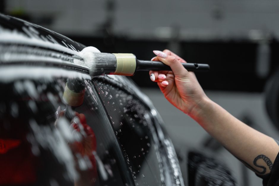 A close-up of a person's hand holding a large cleaning brush with a foam head, using it to clean the exterior windshield and front hood of a black car inside a garage or loading area. The person has well-manicured nails and a small tattoo on their wrist. The background shows part of a vehicle or equipment in the electronics, with the focus primarily on the cleaning action and tools involved in the house removal or moving process. This scene illustrates cleaning tasks related to home relocation, such as preparing furniture or vehicles for transport, supported by the professional services of Man with Van East Sheen, specialising in removals and packing for house clearance or move-in readiness.