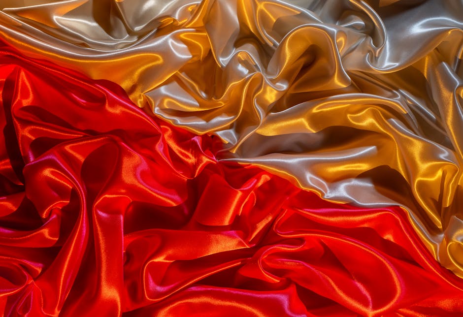 Close-up image of crumpled satin fabric in vibrant red, gold, and silver colours, with the shiny material reflecting light and creating highlights and shadows that accentuate the smooth, flowing texture. The fabric appears to be used in packing or wrapping materials during home relocation or furniture transport, with folds and creases indicating it has been folded or bundled. The setting suggests an indoor environment possibly associated with moving preparations or packing activities, relevant to house removals services like those offered by Man with Van East Sheen. The detailed surface textures and vivid colours emphasize the importance of protective wrapping in the process of packing and moving belongings carefully.