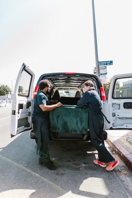 Two professional movers from Man with Van East Sheen are loading a large, dark green upholstered furniture piece into the rear of a white cargo van parked on a street in East Sheen. The interior of the van is visible with its open doors, revealing padding materials and space for furniture transport. One mover, wearing dark clothing and a blue shirt, is holding the furniture from one side, while the other, dressed in a dark jacket and red shoes, is guiding it from the opposite side. The scene is set outdoors during the daytime, with a clear sky and a street sign reading 'Somerset' visible in the background, along with other parked vehicles and urban surroundings. This image demonstrates the careful handling and professional loading process involved in household relocations, emphasizing the importance of proper packing and vehicle organisation for efficient furniture transport during home removal services offered by Man with Van East Sheen.