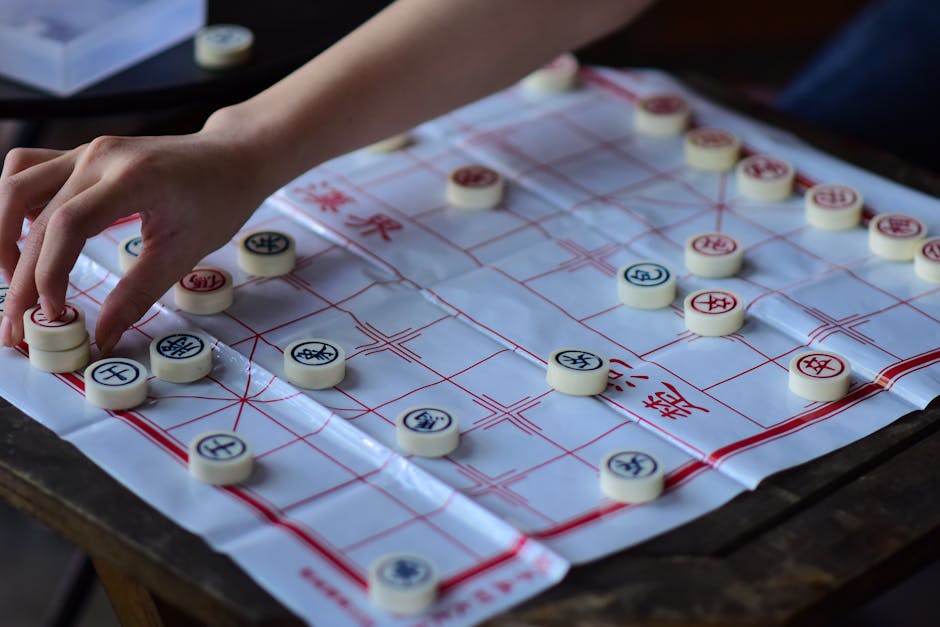 A close-up view of a traditional Chinese chess set arranged on a plastic game board with red grid lines and Chinese characters, placed on a dark wooden surface outdoors. A person's hand is seen moving a white game piece with black Chinese characters on it, while other game pieces, mostly white with red or black Chinese characters, are positioned on the board. Surrounding the board, there are no additional objects visible, but the scene suggests an outdoor environment suitable for casual play or a break from home relocation activities. The lighting is natural, highlighting the details of the game pieces and the board, and the scene emphasizes a moment of strategic move during a Chinese chess game.