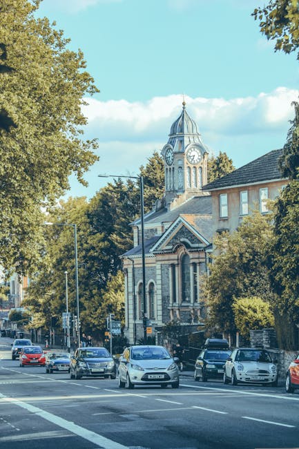 A street scene in East Sheen featuring several parked cars alongside a sidewalk with a large historic church building in the background, identifiable by its clock tower with a pointed spire and ornate architectural details. The image is taken during daylight hours with clear blue skies and some scattered clouds, surrounded by leafy trees providing partial shade. The foreground shows the edge of the roadway and pavement, suggesting an urban environment suitable for home relocation and delivery services. This scene visually supports the context of local removals and furniture transport, as offered by Man with Van East Sheen, highlighting the typical setting for loading or unloading furniture and boxes during a house move or packing and moving operation.