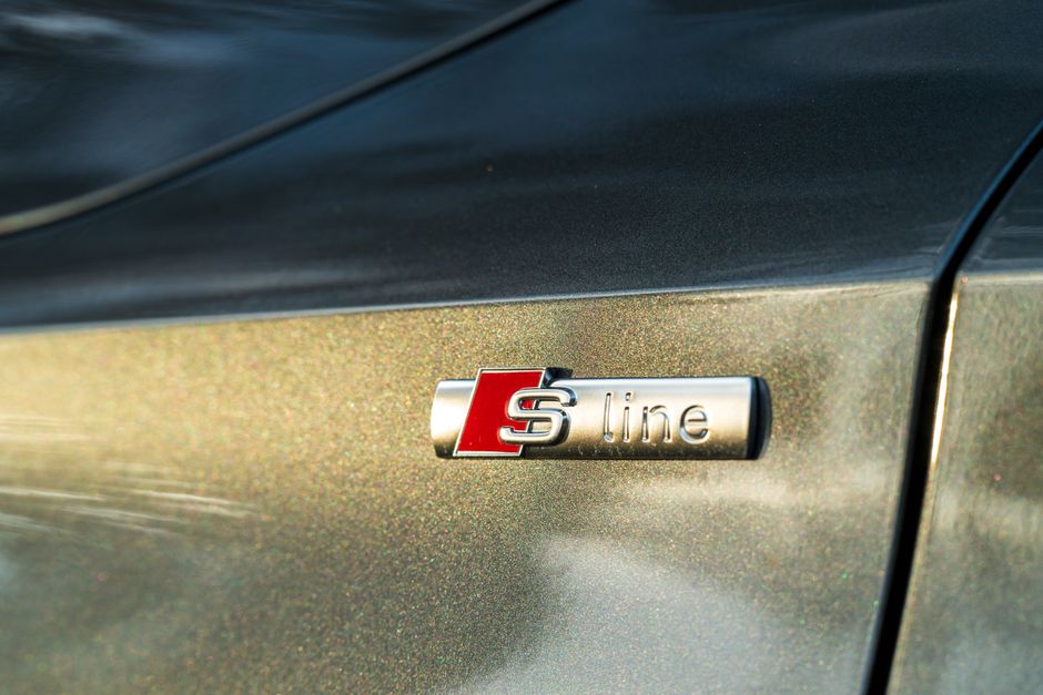Close-up of a metallic emblem on a dark car door displaying the 'S line' badge, indicating a sporty trim of the vehicle, with a smooth reflective surface and subtle metallic flakes visible in the paint. The image captures the badge positioned just above a seam where the car panel meets, with a blurred background suggesting the vehicle is stationary outdoors. The detailed view focuses on the emblem's design, highlighting the contrast between the metallic lettering and the dark, glossy paint finish, with sunlight reflecting subtly on the surface. This image may relate to vehicle transportation or packing for a home relocation involving a car, as part of relocating services offered by Man with Van East Sheen, though only the badge and part of the vehicle's body are visible, emphasizing the vehicle's condition and branding.