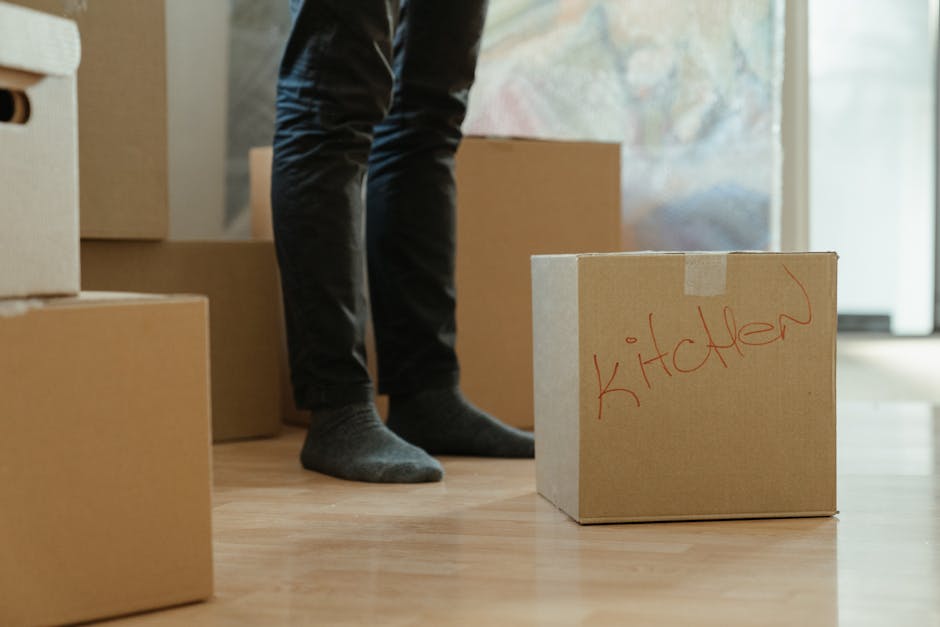 Inside a home with natural daylight coming through a window, a person is preparing for a house removal. The individual is wearing dark trousers and grey socks, and is seen lifting or positioning a cardboard box labeled 'kitchen' with red marker. Several other cardboard boxes are nearby, some stacked, indicating packing in progress. The floor is wooden, and the boxes are of various sizes, made from plain brown cardboard, suitable for packing kitchen items or household belongings. In the background, part of the doorway and an open living space are visible, suggesting a move or packing process within the property. The scene captures the early stages of a home relocation, with furniture or appliances yet to be moved, and hints at the involvement of professional movers such as Man with Van East Sheen, who handle packing and furniture transport as part of their removals services.