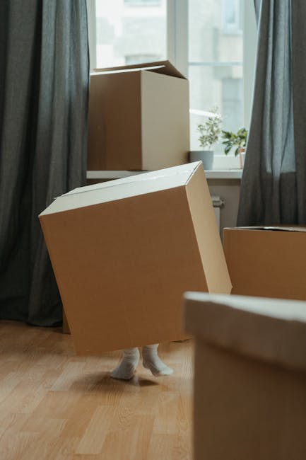 Inside a home with natural daylight coming through a window, a person is preparing for a house removal. The individual is wearing dark trousers and grey socks, and is seen lifting or positioning a cardboard box labeled 'kitchen' with red marker. Several other cardboard boxes are nearby, some stacked, indicating packing in progress. The floor is wooden, and the boxes are of various sizes, made from plain brown cardboard, suitable for packing kitchen items or household belongings. In the background, part of the doorway and an open living space are visible, suggesting a move or packing process within the property. The scene captures the early stages of a home relocation, with furniture or appliances yet to be moved, and hints at the involvement of professional movers such as Man with Van East Sheen, who handle packing and furniture transport as part of their removals services.